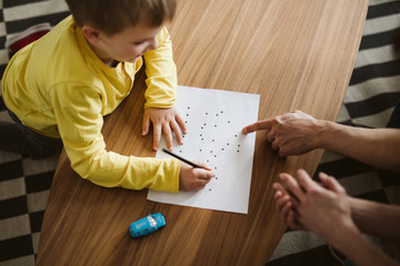 Cute boy kneeling on the floor and connecting dots on a piece of paper - obrazy, fototapety, plakaty