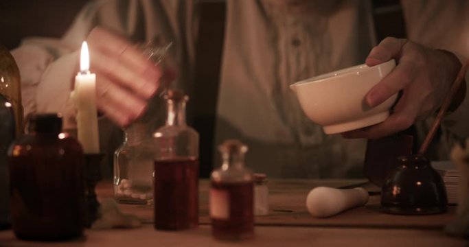 A man skilled in apothecary pours a powdered mixture from a mortar and pestle into a solution then swirls the concoction.