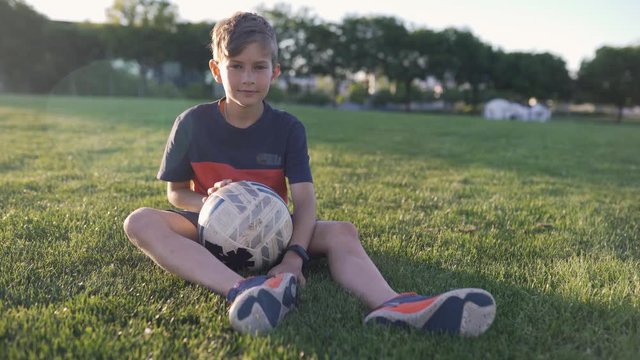 portrait of a boy football player with a ball in a park sitting on the lawn looking at the camera and smiling overall plan