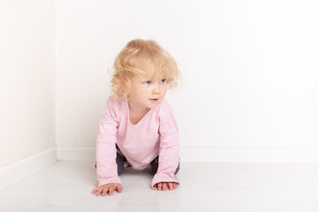 curly blonde baby girl in comfortable home casual clothes is lying on the floor in a bright white room smiling and looking at the camera