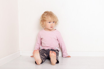 Naklejka premium curly blonde baby girl in comfortable home casual clothes is lying on the floor in a bright white room smiling and looking at the camera
