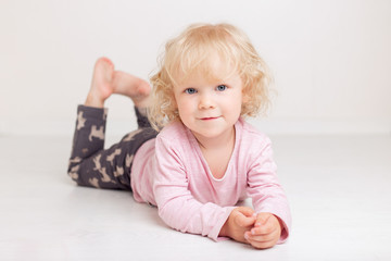 curly blonde baby girl in comfortable home casual clothes is lying on the floor in a bright white room smiling and looking at the camera