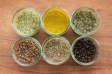 Six glass jars with black pepper, allspice, coriander, caraway, dried basil, oregano and curry on a brown wooden cutting board. Spices top view.