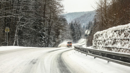 Snow covered forest road, ice on asphalt, orange gritter plough truck coming in distance, dangerous...