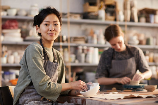 Portrait of content attractive young Asian female artisan in apron sitting at desk and polishing ceramic bowl with sandpaper in crockery workshop