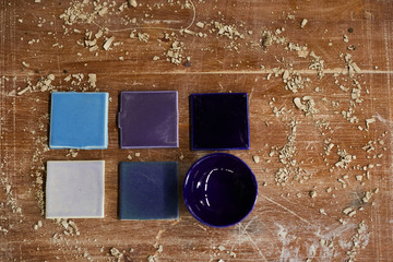 High angle view of ceramic samples in blue shadows and purple bowl placed on dirty scratched table with clay dust