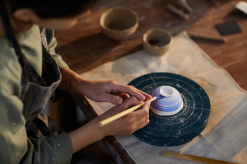 Close-up of concentrated female potter in apron sitting at table and painting ceramic bowl on stand, copy space