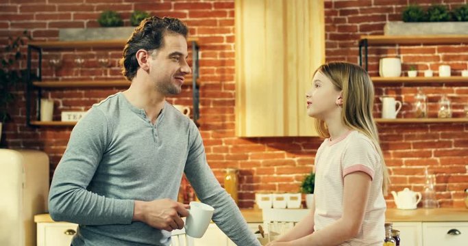 Happy handsome Caucasian father standing with a coffee or tea in hand and talking cheerfully with his pretty cute teen daughter.
