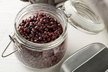 Raw, uncooked, dried adzuki (red mung) beans in glass storing jar with metal spoon on white wood table background