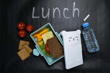 Obraz premium open lunch box with a sandwich of whole grain bread, cheese, green salad, tomato, cucumber and a bottle of water on a black background with colored crayons and lunch inscription written in white chalk