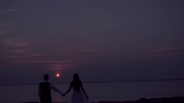 loving couple - young groom and beautiful bride at sunset , silhouette view. They are holding hads