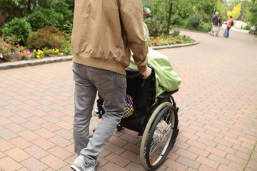 Obraz premium A man in gray jeans carries an elderly woman, a disabled person in a wheelchair on the street for a walk, rear view