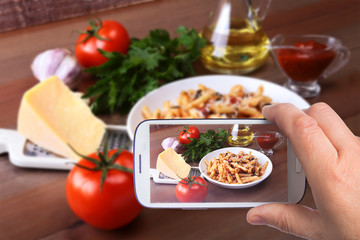 hands of man with smartphone taking photo Spicy penne pasta bolognese with vegetables, chili and cheese in tomato sauce on the background of tomatoes, garlic, olive oil and spices. Top view.