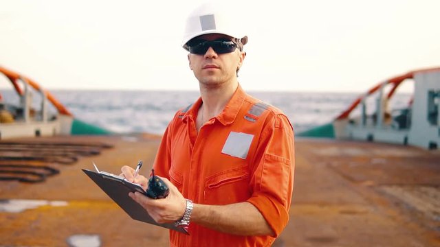 Marine chief officer or chief mate on deck of ship or vessel. He fills up ahts vessel checklist. Ship routine paperwork. He holds VHF walkie-talkie radio in hands.