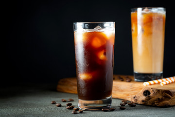 Ice coffee in a tall glass with cream poured over, ice cubes and beans on a old rustic wooden table. Cold summer drink with tubes on a black background with copy space
