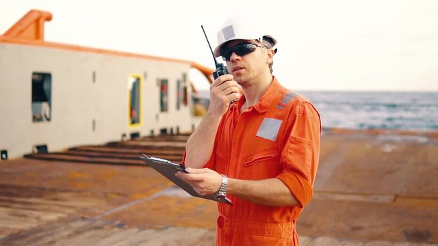 Marine chief officer or chief mate on deck of ship or vessel. He fills up ahts vessel checklist. Ship routine paperwork. He holds VHF walkie-talkie radio in hands.