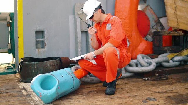 Marine chief officer or chief mate on deck of ship or vessel. He fills up ahts vessel checklist. Ship routine paperwork. He holds VHF walkie-talkie radio in hands.