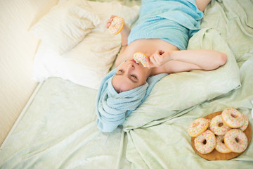The girl in a towel has breakfast in the bed. A young attractive woman eats donuts in bed. The girl with a towel on her head happily eats donuts.