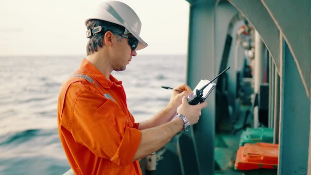 Marine chief officer or chief mate on deck of ship or vessel. He fills up ahts vessel checklist. Ship routine paperwork. He holds VHF walkie-talkie radio in hands.