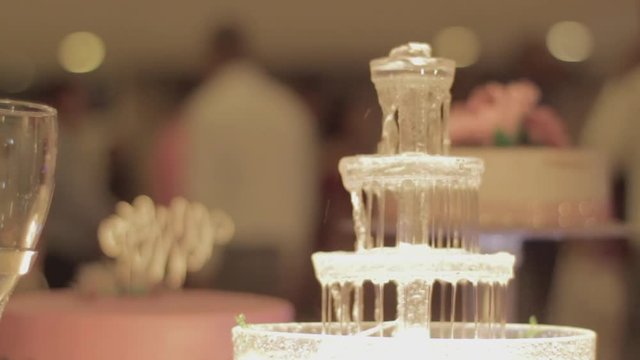 Focus pull from blurred wedding reception scene to close up on crystal glass table setting fountain alongside champagne flutes as blurred people walk in background.