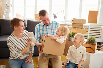 Fototapeta na wymiar Cheerful positive young parents entertaining younger kid into new home: father holding cardboard box with little daughter while mother showing toy to her