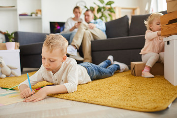 Serious concentrated elder son in casual outfit lying on fluffy carpet and drawing with pencils...