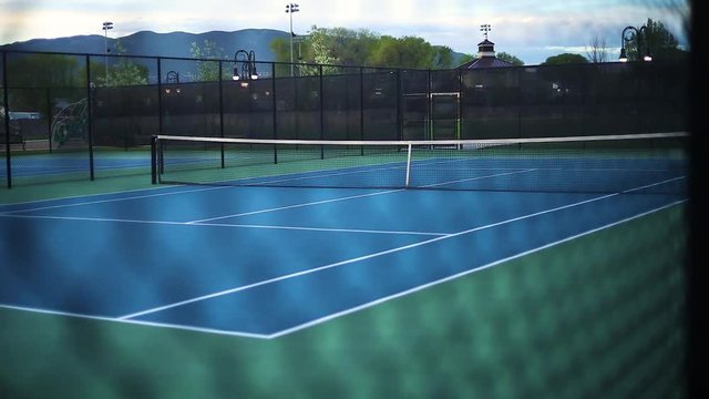 A smooth shot from outside the fence looking in on an empty tennis court. Focuses in on the fence first, then slowly transitions to focusing on the court. A good intro shot.