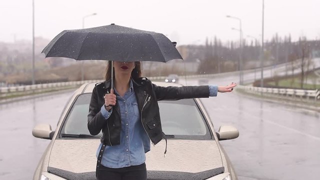 A young upset girl with an umbrella catches a car standing on the road in the rain with snow, her car ran out of gas and she asks for help.