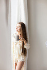 Attractive young girl in night clothes standing by the window with a mug of coffee