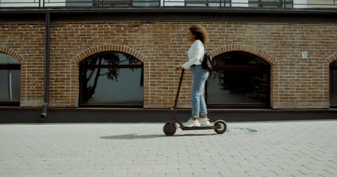 SIDE VIEW Attractive African American black female riding an electric city shared scooter in the street. 4K UHD 60 FPS SLOW MOTION