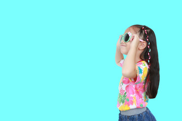 little Asian kid girl wearing a flowers summer dress and sunglasses looking up isolated on cyan background with copy space. Summer and fashion concept. Side view.
