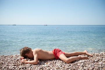 Fototapeta na wymiar tired little boy in red shorts lying on stone beach near ocean sunbathe during summer holiday travel activity with copy space