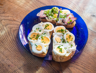 close up fresh home made rye bread sandwich with egg slices, ham bacon, chive and mayonnaise on blue plate on wooden table