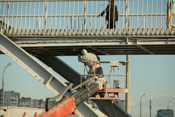 Naklejka premium Professional worker in a protective suit and mask paints a bridge from the spray gun stand on the hydraulic lift.
