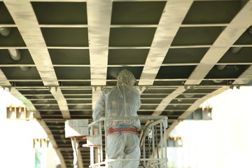 Fototapeta premium Professional worker in a protective suit and mask paints a bridge from the spray gun stand on the hydraulic lift.