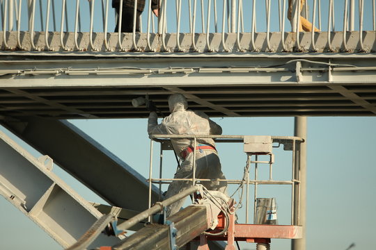 Professional worker in a protective suit and mask paints a bridge from the spray gun  stand on the hydraulic lift.