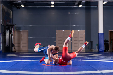 Two strong wrestlers in blue and red wrestling tights are wrestlng and making a  making a hip throw  on a yellow wrestling carpet in the gym. Young man doing grapple.