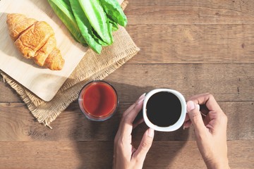 Top view woman hands holding black coffee cup, tomato juice, croissant and vegetable on rustic wooden table