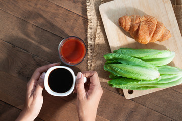 Creative flat lay of breakfast concept. Woman hands holding coffee cup with tomato juice, croissant and vegetable on wooden table