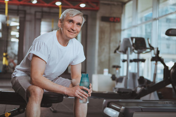 Cheerful senior man relaxing after working out at the gym, holding bottle of water, smiling joyfully. Happy healthy elderly man resting after exercising at sport studio, drinking water. Retirement, li