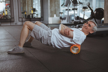 Cheerful senior man smiling to the camera, using foam roller on his back. Elderly sportsman relaxing his back muscles after working out at the gym. Massage, medical, health concept