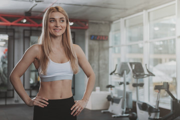 Charming blond haired young fitness woman in sports top and leggings posing confidently after working out. Cheerful female athlete enjoying morning workout at sport studio, copy space