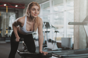 Fototapeta premium Happy beautiful female athlete smiling, doing triceps exercise at sport studio, copy space. Lovely fitness woman enjoying her morning workout at the gym, lifting dumbbells