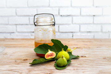 Antique glass preserve bottle with fig preserve together with a fresh green fig leaf and halved ripe figs on a raw wood surface. 