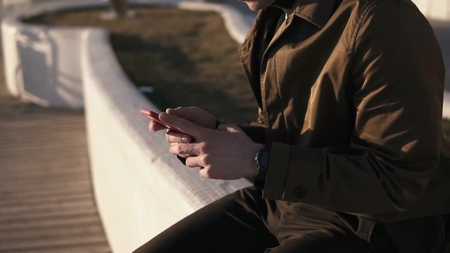 Side view of a elegant nab in glasses and brown coat sitting outside on parapet near the seaside. Brunette young man looking at his smarphone, texting. Sunny, windy cool weather