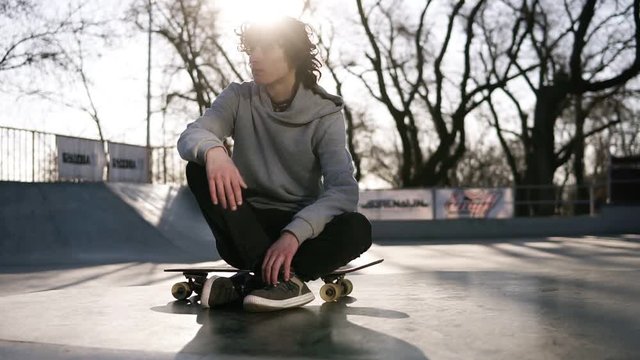 Skater in light grey hoodie sitting on his board looking thoughtful outside the skate park. Sun strongly shines just above his head