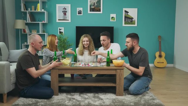 Company of boys and girls playing board game in living room and drinking beer