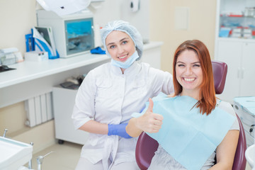 Obraz premium A female dentist and a red-haired patient in an office after dental treatment are looking at the camera with happy faces. Happy woman with her doctor after taking shows sign for shelter.