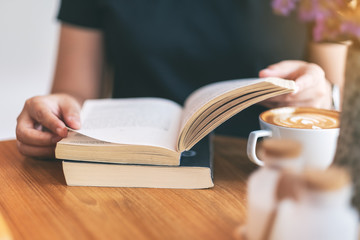 Closeup image of a woman holding and reading a vintage novel book with coffee cup on wooden table