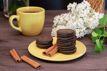Tea pair cup and yellow saucer. Chocolate chip cookies, cinnamon sticks, white lilac on a dark wooden table